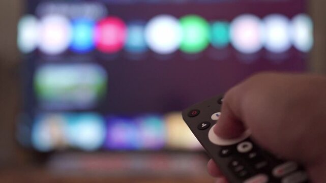 hand holding TV remote control, browsing streaming apps on blurred smart television screen, home entertainment technology concept.