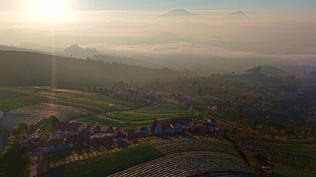 Drone shot of tropical highland farmland covered in morning mist, traditional village houses, rolling hills and volcanic mountains in the background.