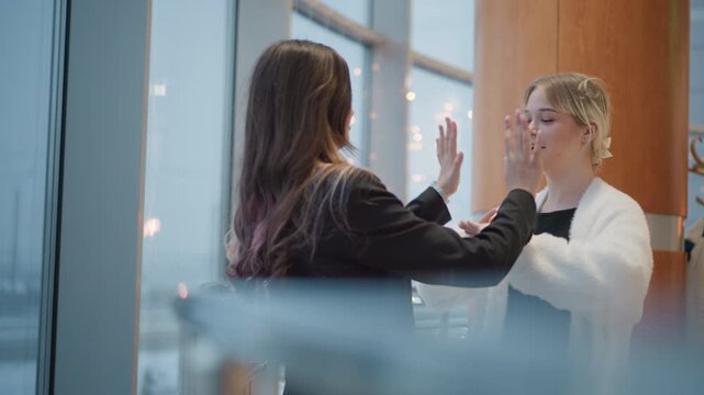 Two young women stand beside floor-to-ceiling window in bright office space, smiling and talking closely while one touches other s arm and chest in warm supportive exchange, with women, support