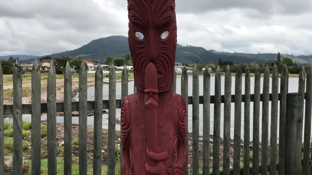 Red maori pouwhenua carving with paua shell eyes standing guard at the historic Ohinemutu village, revealing the settlement and St. Faith's Anglican Church in Rotorua, New Zealand
