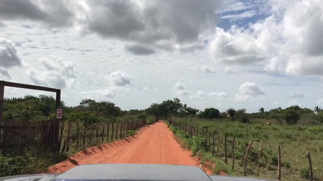 First-person perspective from an off-road vehicle driving along a bumpy red sand road in rural Jericoacoara, Brazil, through lush green vegetation under a cloudy sky on a sunny day