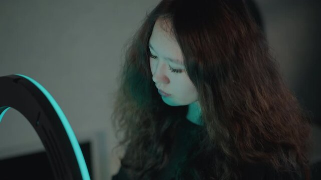Young woman with long curly hair leans toward illuminated ring light in moody indoor setting, gazing downward with contemplative focus, soft purple blue light and calm grooming mood