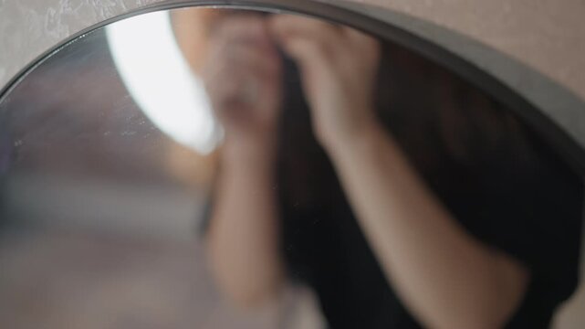 Young woman applies eyeliner while looking into round mirror, steadying eye with fingers and guiding cosmetic tool along upper lash line under soft ring light in warm indoor setting