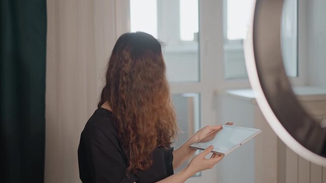 Woman with long curly hair stands indoors near bright window, holding tablet at chest level and angling screen toward camera in clear presenting gesture, with face partly covered by hair