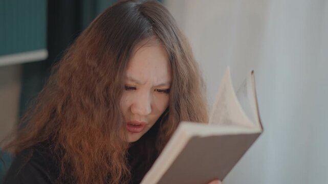 Close frontal view of young woman with long wavy brown hair leaning forward while reading paperback book near bright window, showing puzzled frown, focused gaze, and mild frustration