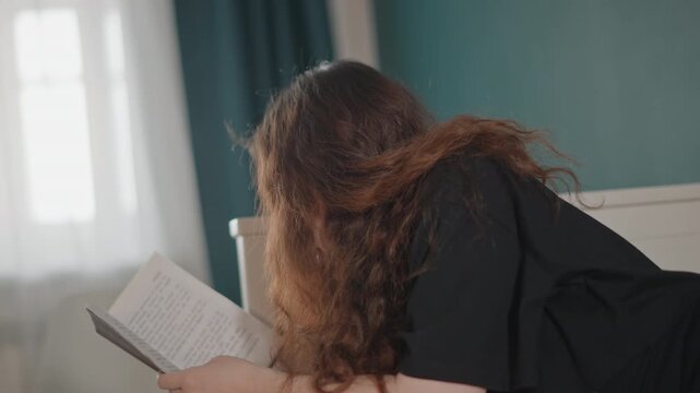 Young woman on stomach across bed, leaning on elbows while reading open book; long curly hair falling forward, page turning, soft window light, calm cozy bedroom scene with quiet domestic comfort