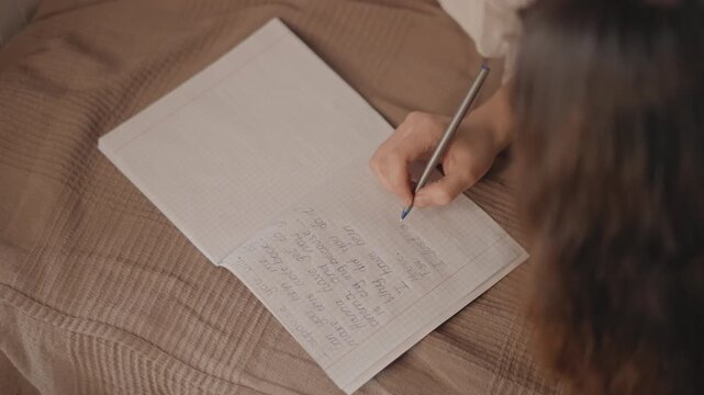 Oblique overhead close-up shows hand holding pen and actively writing cursive notes in open notebook resting on textured brown-beige blanket, with soft warm light and slight motion blur