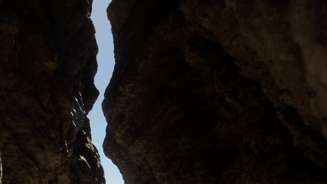 Two large rock walls rise towards the sky with a narrow gap in between. The sun shines bright overhead on a clear day. This canyon is part of a natural landscape.