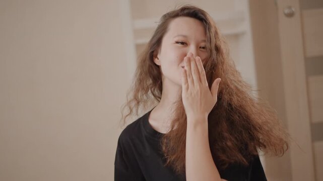 Young woman with long curly hair performs playful grooming gestures and affectionate poses indoors, running hand through hair, touching face, flipping hair toward camera, and forming heart with