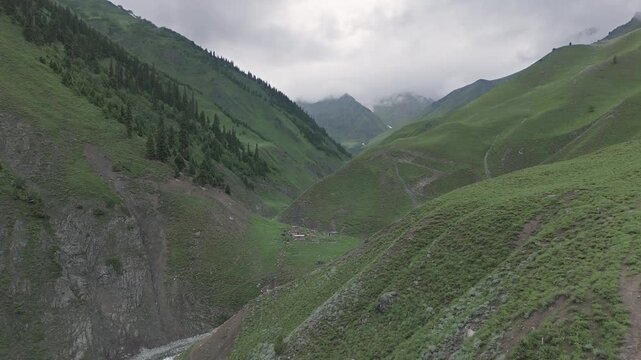 Alpine landscape with a small rural village nestled in the foothills under a cloudy sky, Aerial drone video flying forward over a deep green mountain valley towards remote seasonal wooden houses