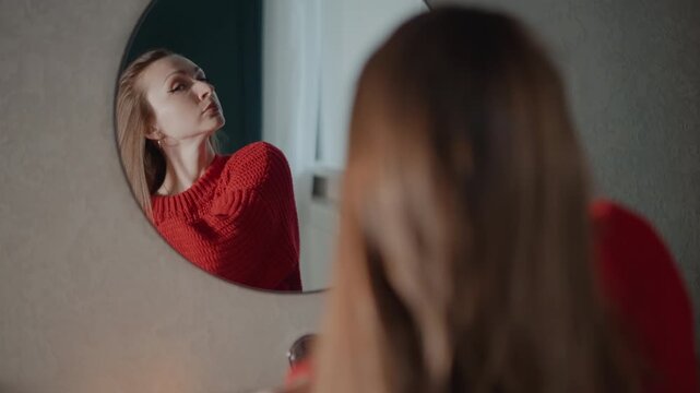 Young woman in red sweater leans toward round mirror, arranging hair, touching earring, and checking face with focused expression in soft indoor light during personal grooming routine