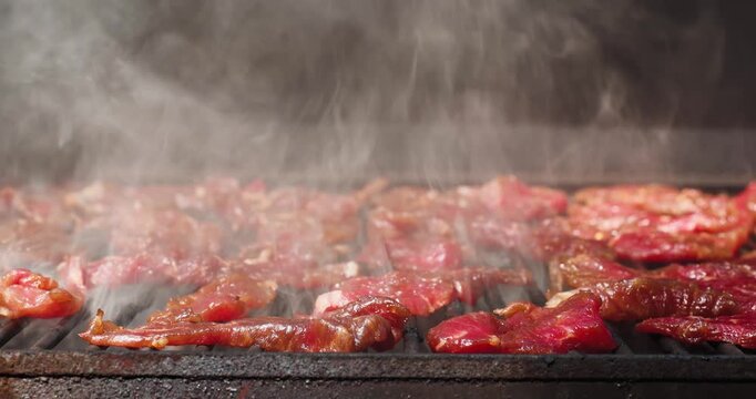 Thin slices of raw round steak slow cook on a pellet grill smoker as steady smoke rises across the grates. Homemade beef jerky smoking with rich color, texture. Raw round steak.