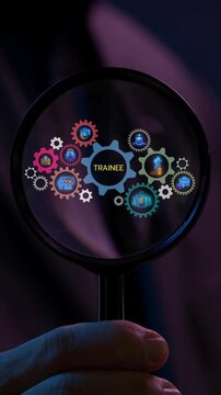 Close-up vertical shot of hand holding magnifying glass to inspect glowing gear mechanism with TRAINEE central text and icons. Corporate internship trainee skill development program audit concept.