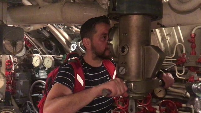 Curious male tourist with a backpack exploring the interior of a historic military submarine, attentively looking through the periscope and rotating it to see the surroundings outside