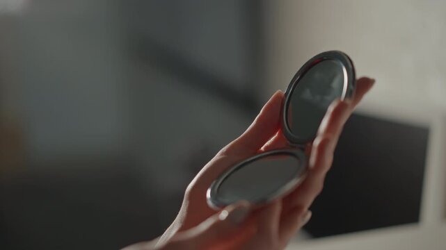 Close-up of hand holding open round compact mirror tilted upward with fingers and thumb supporting case, slight wrist rotation indicating active inspection and makeup adjustment under soft indoor