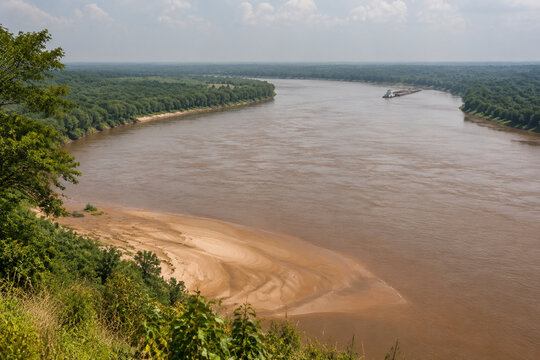 View from high bluff of wide brown river bending through low green forest, sandbar, tugboat and barge downstream, Mississippi River landscape