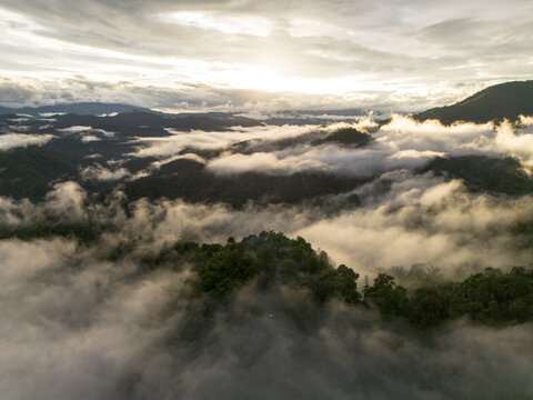 Aerial mountain landscape, cloud, forest, mist, and sunrise create dramatic natural panorama above remote green valley, with layered ridges fading beneath soft atmospheric haze