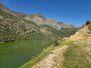 Obraz premium A scenic spring river flowing through rugged green mountains, with a dirt road along the hillside beneath a clear blue sky. 📍 Hawraman Takht, Kurdistan, Iran 