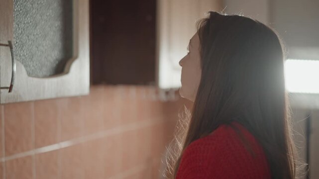 Waist-up view shows young woman in red long sleeve top standing by bathroom mirror, gently touching lips while checking makeup, with warm tile backdrop, soft light, and quiet home routine