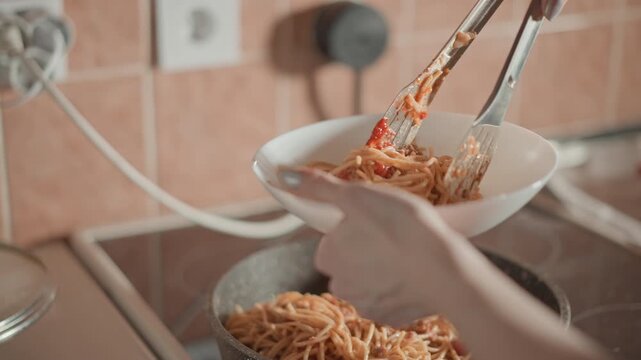 Close-up of hands using stainless tongs to lift and transfer glossy, steaming spaghetti from skillet into white ceramic bowl above stovetop; noodles dangling and falling show active serving motion