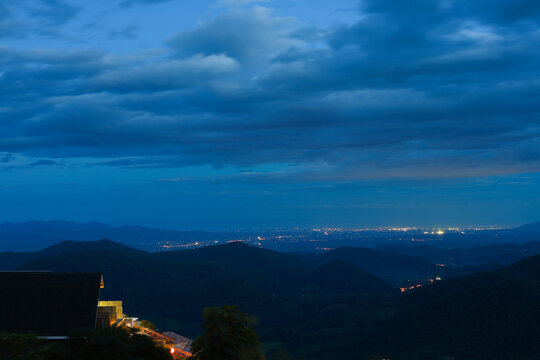 Night mountain landscape with distant city light beneath dramatic blue cloud and illuminated hillside building, creating quiet atmospheric travel scene with layered forest ridge