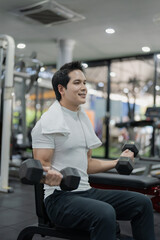 Fototapeta premium Asian man sitting on workout bench doing bicep curls with dumbbells in gym