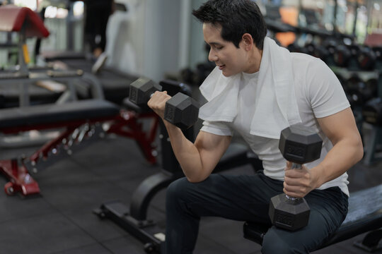 Asian man sitting on workout bench doing bicep curls with dumbbells in gym
