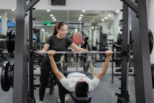 Asian man lifting barbell on bench press with female personal trainer supervising in gym