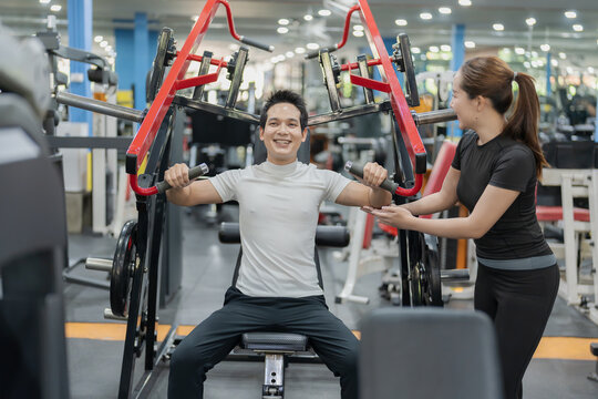 Asian man lifting barbell on bench press with female personal trainer supervising in gym