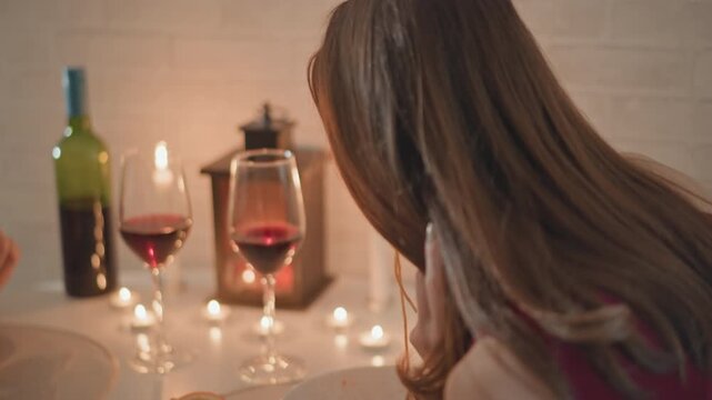 Woman turns and reaches toward food at candlelit dining table, showing visible arm movement and relaxed profile in warm indoor setting with wine glasses, breadboard, soft reflections and intimate mood