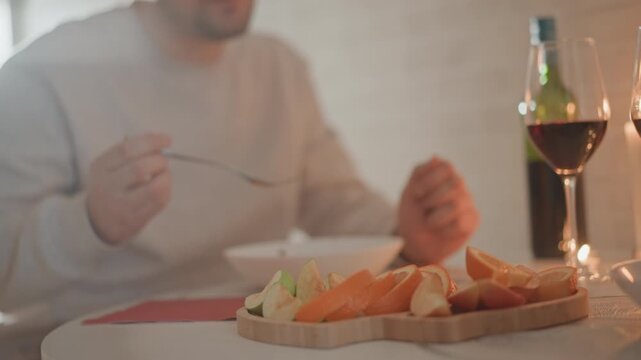 Seated person reaches with fork toward wooden serving board of sliced bread and carrot sticks as wine glasses and bottle rest nearby, with soft warm light and gentle hand motion, with hand, reaching