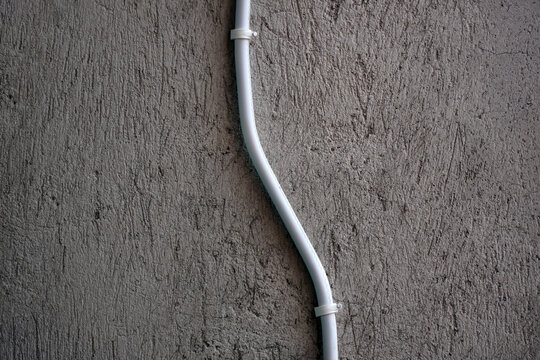 White curved electrical conduit pipe secured with clamps on a rough textured grey concrete wall. Minimalist industrial design, home renovation, electrical installation, and wiring concepts.
