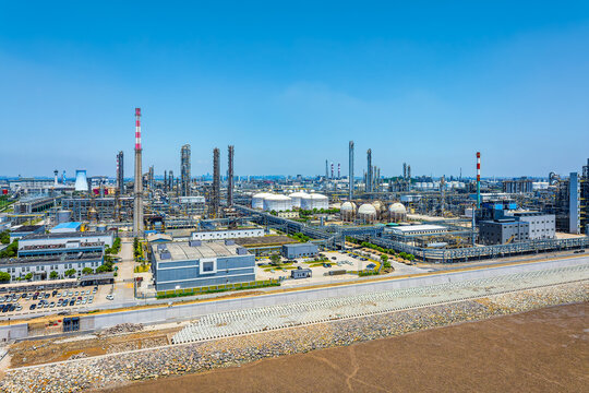 Coastal petroleum refinery with distillation towers, storage tanks, pipelines, and seawall under clear blue sky, illustrating energy infrastructure and industrial development.