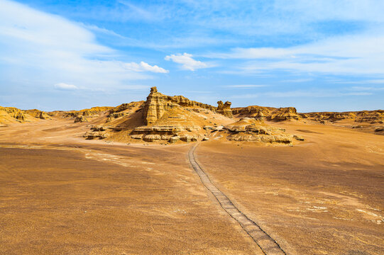 Golden yardang formations rise from the barren Dahaidao desert in Xinjiang, China, with a worn trail across an eroded landscape under a blue daytime sky.