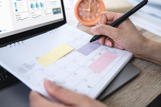Close up of person planning schedule, writing notes on calendar planner with colorful sticky notes and laptop displaying financial analytics.