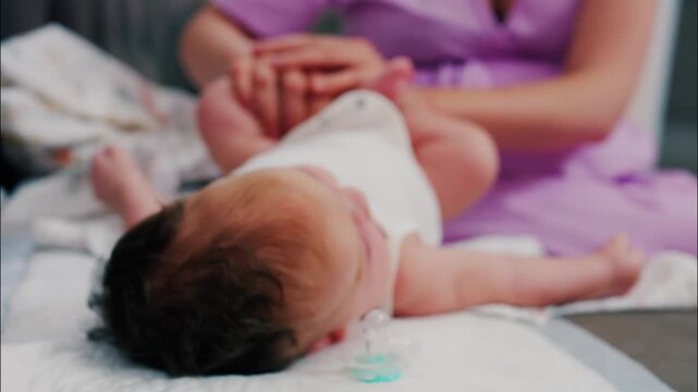 Newborn baby on changing table with pacifier nearby, mother gently massages childs feet in soft focus background.