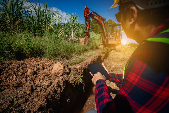 Dedicated Asian engineers in brightly colored work uniforms are monitoring digital data on tablets at a construction site with exposed pipes, sewage systems, and technological advancements.
