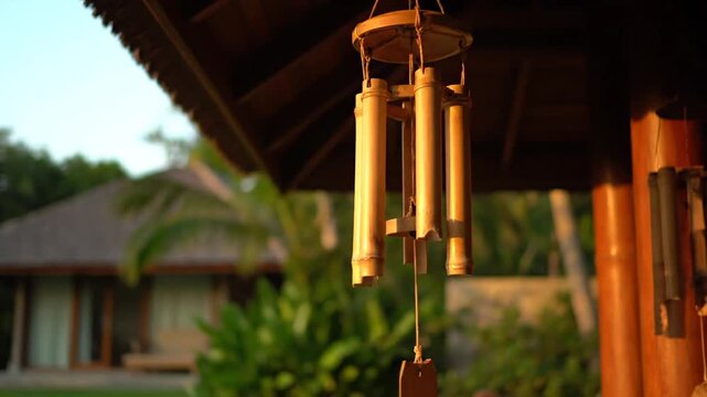 Close-up of a decorative bamboo wind chime gently swaying under a roof in a warm outdoor environment.