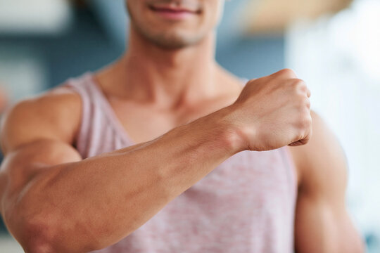 Strong man flexes bicep, showcasing defined muscles. He wears a light-colored tank top. Focus is on his forearm and clenched fist. Confident posture suggests determination or strength