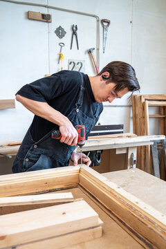Focused woodworker finishing assembling vintage wooden window frame at workplace in workshop, man carpenter using handheld cordless electric drill instrument driving screws in corners of wooden frame.