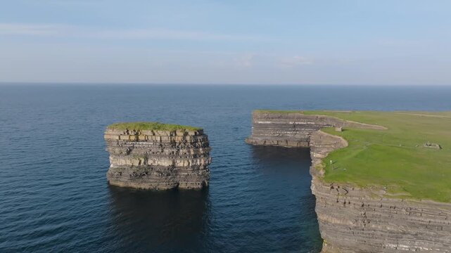 Layered rock sea stack Dun Briste stands beside sheer grassy cliffs under calm daytime skies.