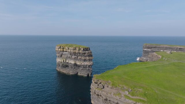Detached Dun Briste sea stack rises beside a sheer green cliff headland over deep blue water on a clear calm day.