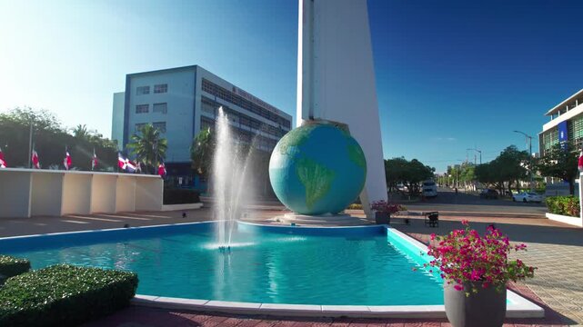 Ground-level aerial tracking shot moving right past a white marble statue, water fountain, and the central globe obelisk monument in Santo Domingo
