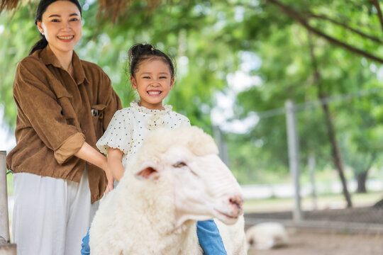 happy preschool child girl riding a sheep with mother beside in outdoor farm