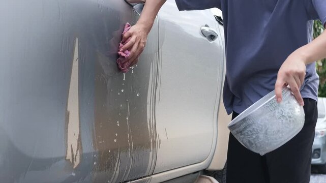 Woman pouring water from a bowl down the dirty side door of a pickup truck then scrubbing off the mud with a cloth