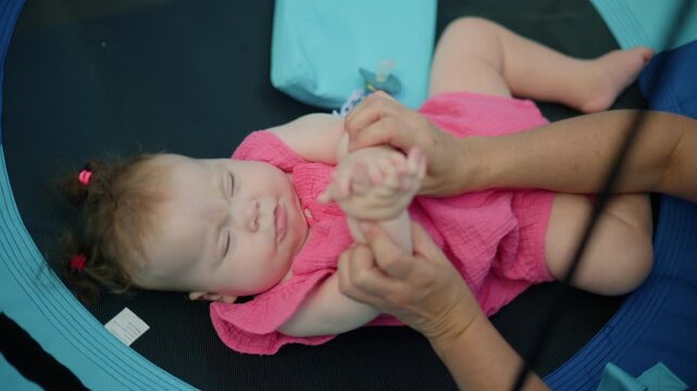 Adult hands gently touching and stimulating a baby wearing a pink dress, lying on a blue sensory swing
