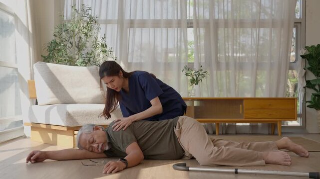Adult daughter giving first aid to unconscious senior man after home accident, walking cane nearby as worried woman responds in bright living room with sofa, plants, and sunlight