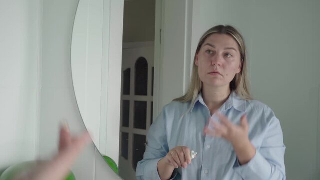 Calm candidate in a light shirt squeezes makeup primer and smooths it onto her cheeks while studying her reflection at home.