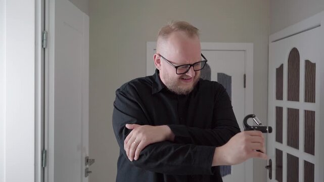 Middle-aged man in black clothing uses a hand tool, then winces and clutches his painful elbow in a bright hallway.