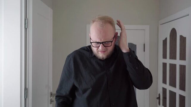 Man in glasses presses and rubs his ear with a strained expression, acting out ear ache or hearing discomfort in a neutral home hallway.